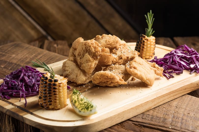 Crispy fried chicken pieces, grilled corn, red cabbage, and a hard-boiled egg on a wooden board.