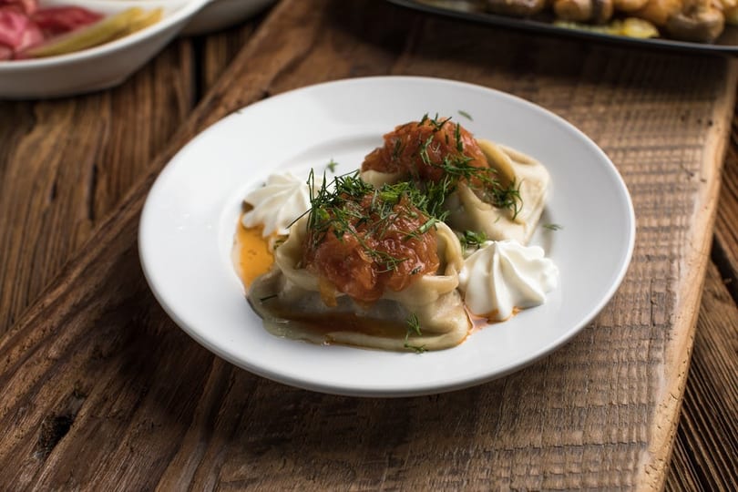 Two dumplings topped with red sauce and dill, garnished with whipped cream on a white plate.