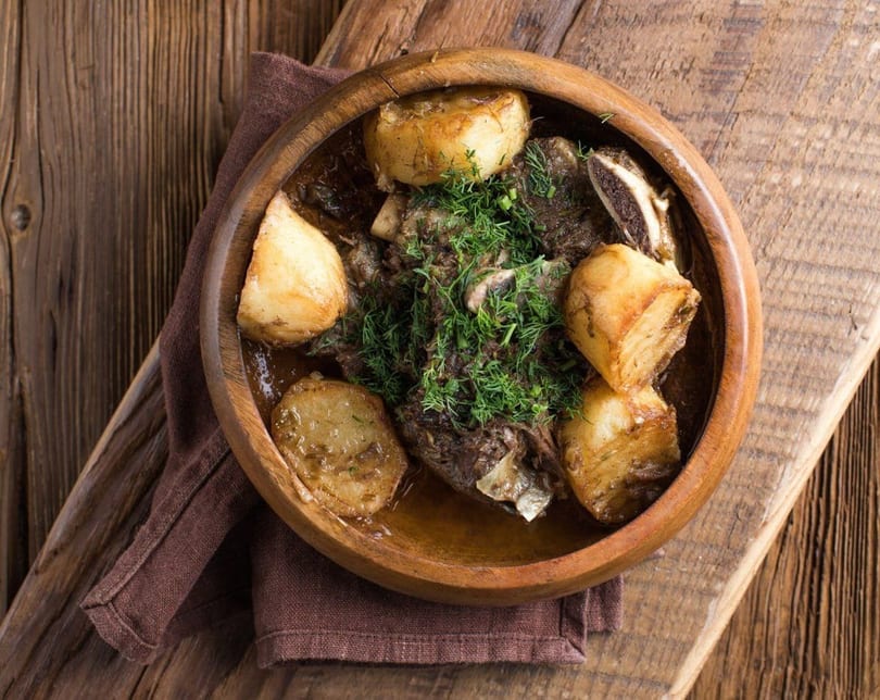 Hearty stew with meat, potatoes, and fresh dill in a wooden bowl on a rustic table.