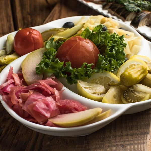 A white platter filled with pickled tomatoes, cucumbers, cabbage, lemons, and green peppers on a wooden table.