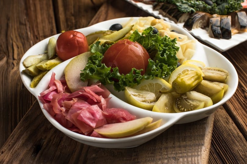 A white platter filled with pickled tomatoes, cucumbers, cabbage, lemons, and green peppers on a wooden table.